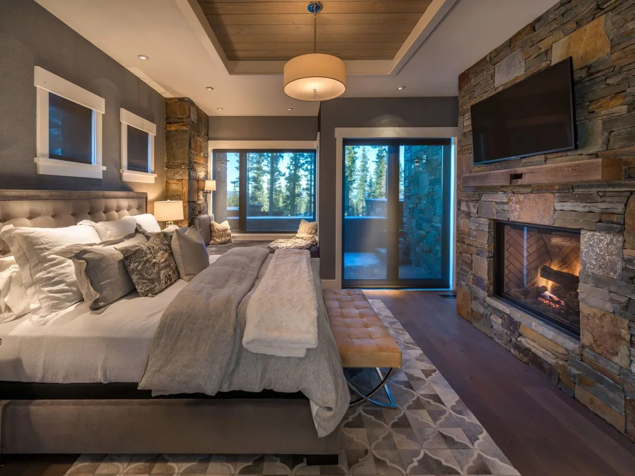 A layered-stone bedroom fireplace with a timber mantel faces a bed beneath a pale timber ceiling inset.