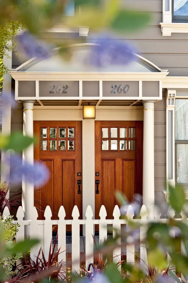 Neighboring wood front doors under shared small canopy with house numbers above and white picket fence