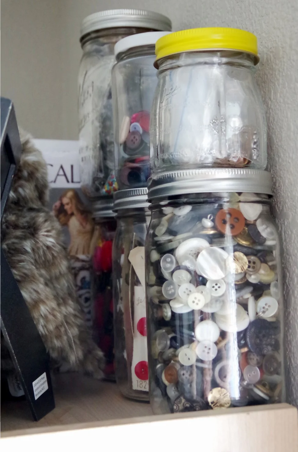 Clear jars with screw-top lids hold buttons and other small sewing supplies on a shelf.