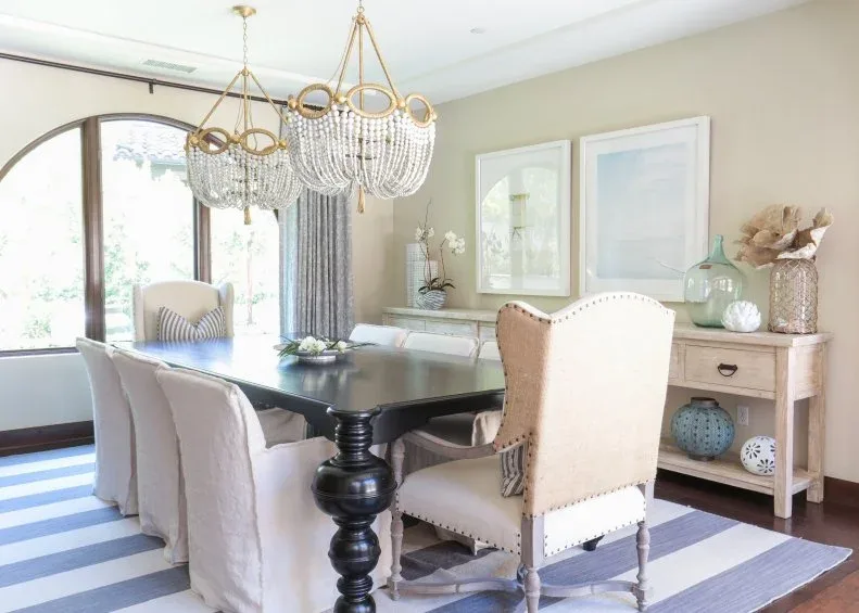 Neutral dining room with dark turned-leg table, cream upholstered chairs, blue striped rug, pale console and beaded chandelier.