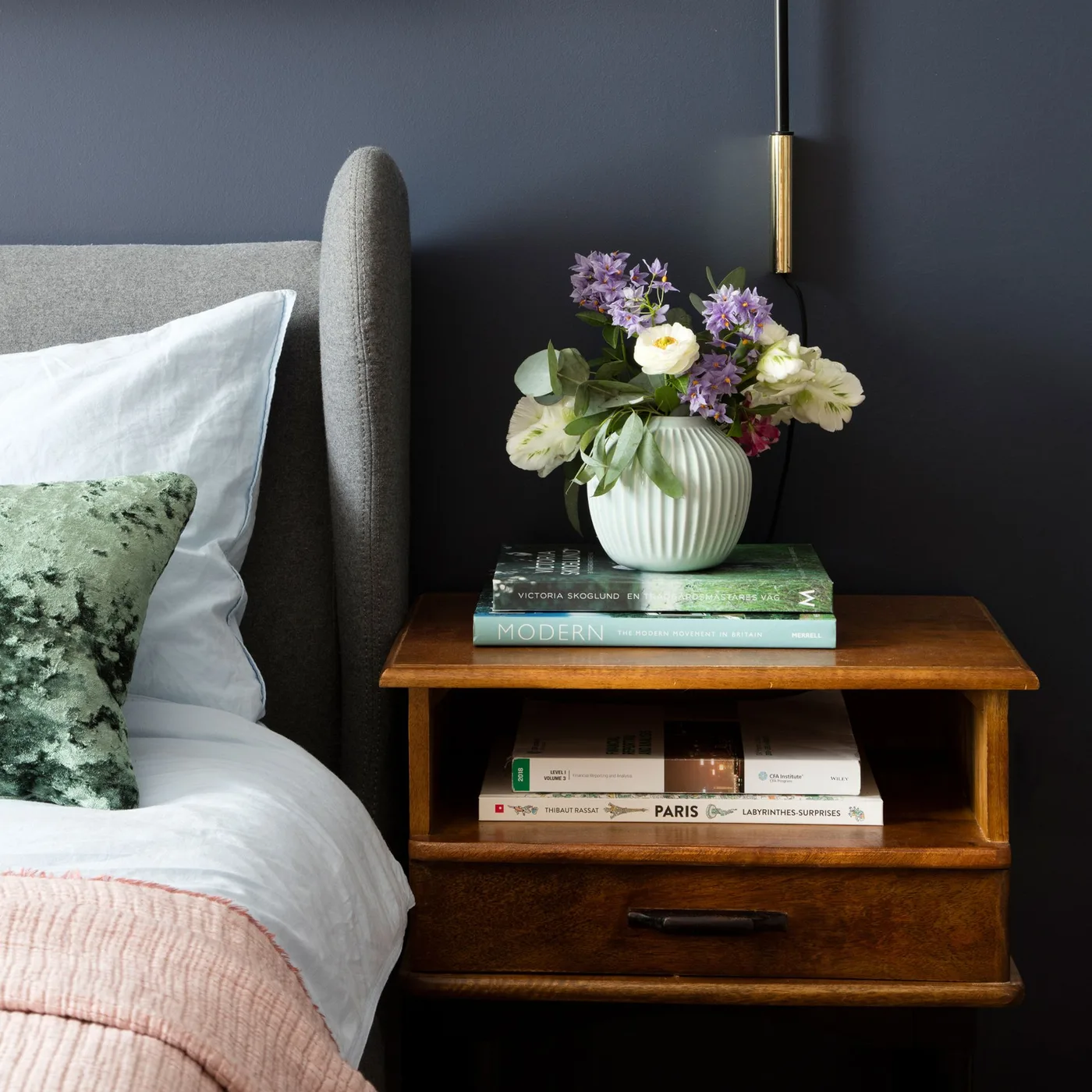 Navy bedroom wall behind a gray winged headboard with a pink bedcover, green patterned cushion, and wooden bedside table.