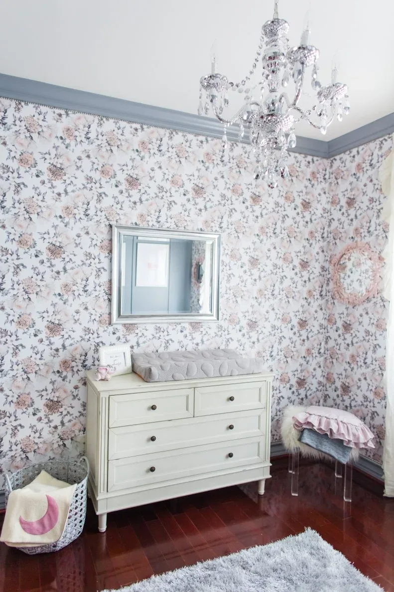 Pale dresser and rectangular mirror against small floral wallpaper with gray trim and decorative chandelier.