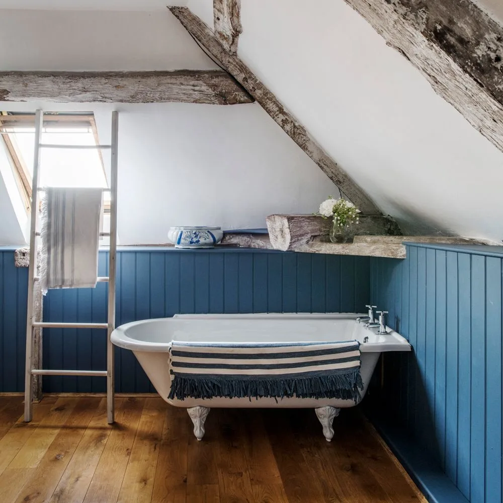Blue lower paneling surrounds a white clawfoot bath beneath exposed roof beams and pale sloping walls