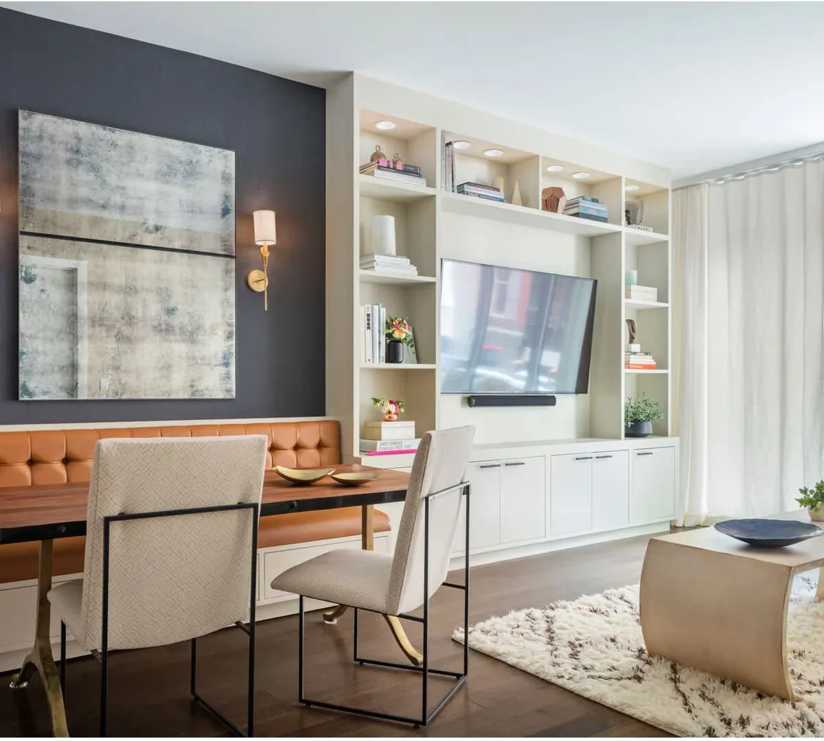 An orange-brown dining bench sits below a dark wall and framed art beside pale built-in television shelves and a light sitting area.