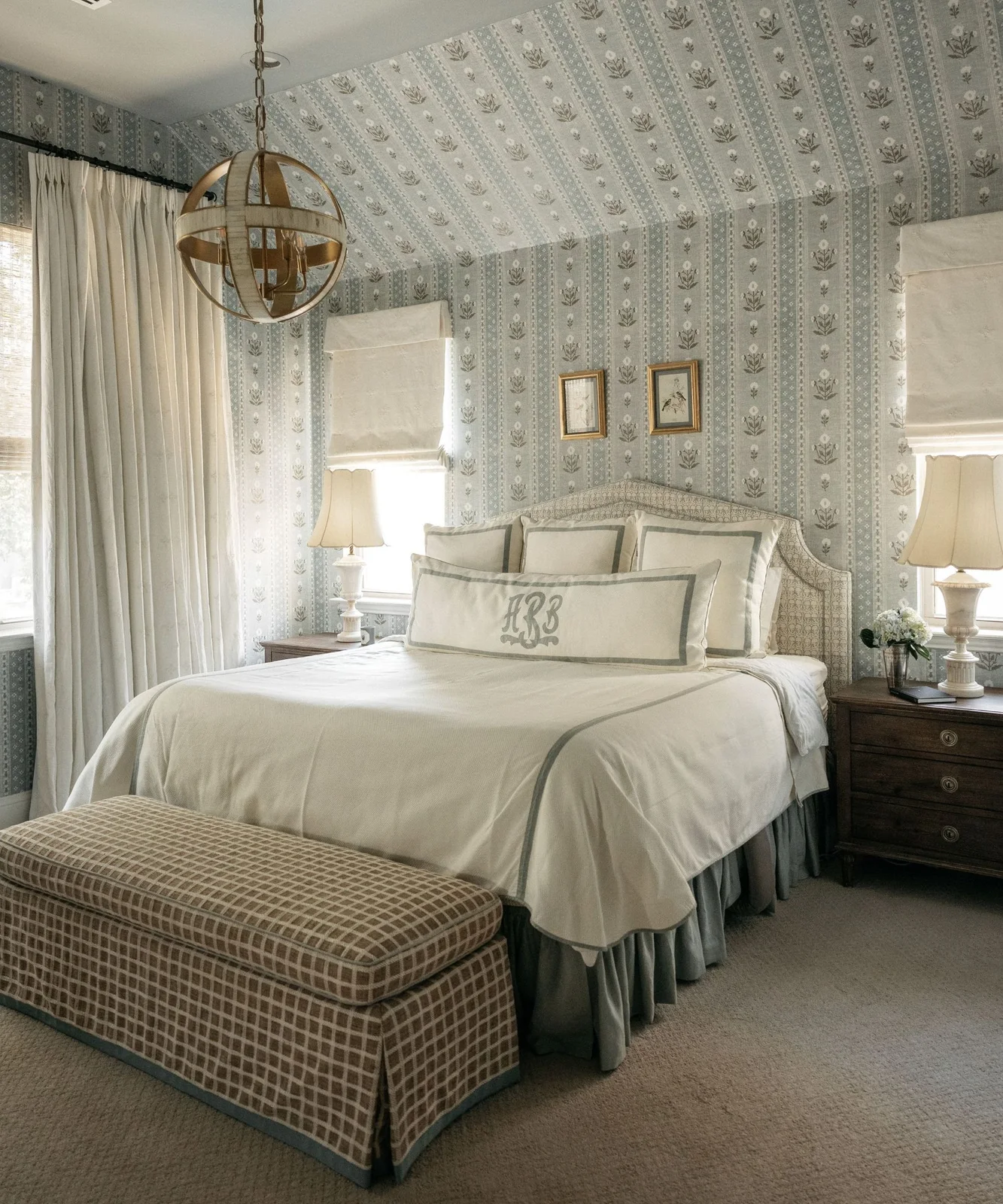 Blue-and-white floral striped wallpaper follows a sloping ceiling above a pale geometric headboard, white bedding and a checked foot bench
