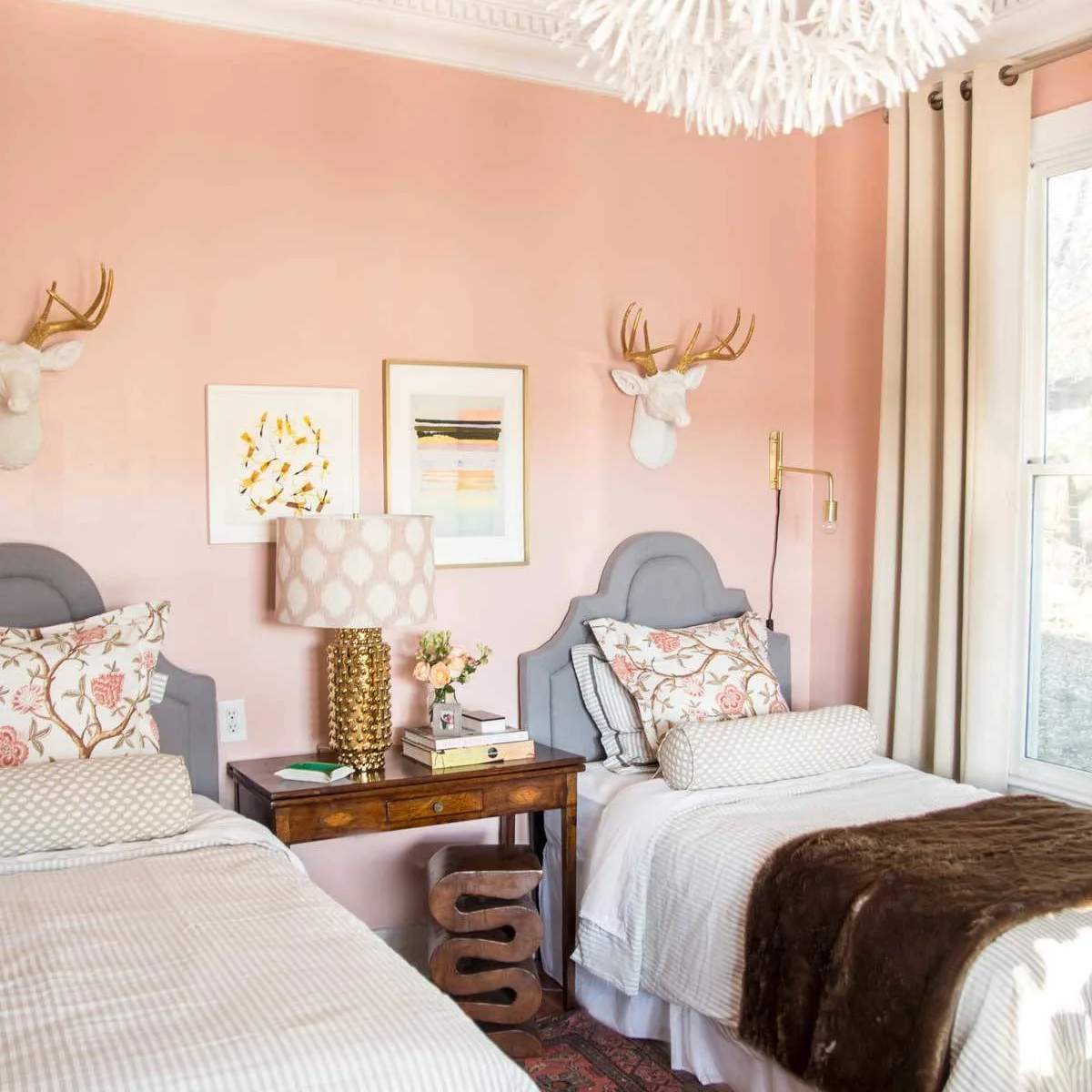Two beds with gray shaped headboards against a peach-pink wall, floral cushions and a shared wood table