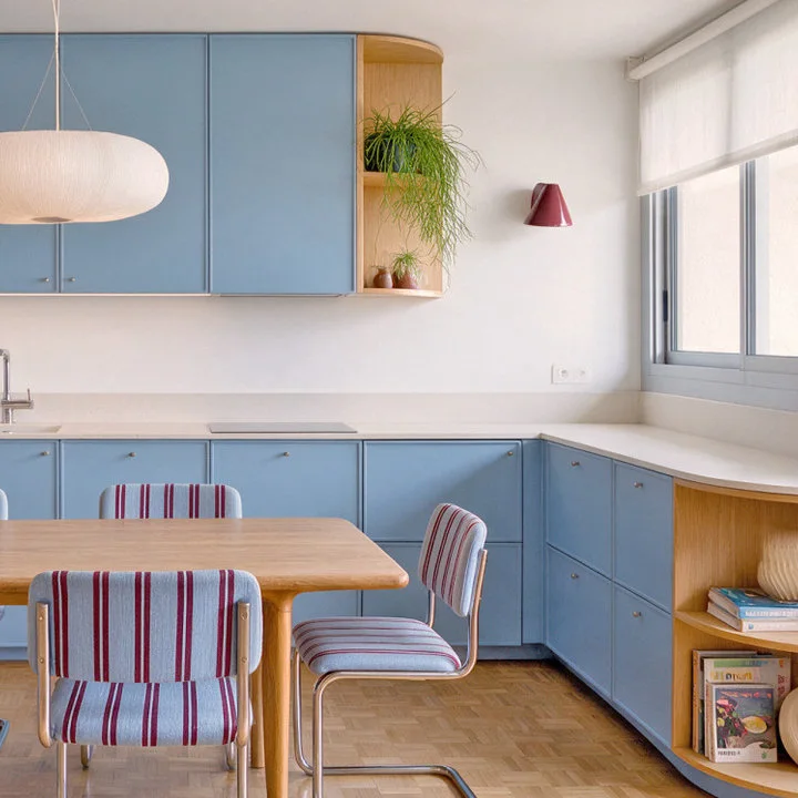 Striped dining chairs and a wood table stand beside pale blue kitchen cabinets, open wood shelves and a white hanging shade.