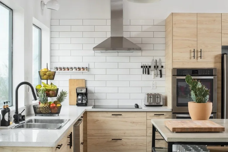 A light wood kitchen has white rectangular wall tiles, dark handles, a metal hood, a double sink and a tall oven cabinet beside a small worktable.