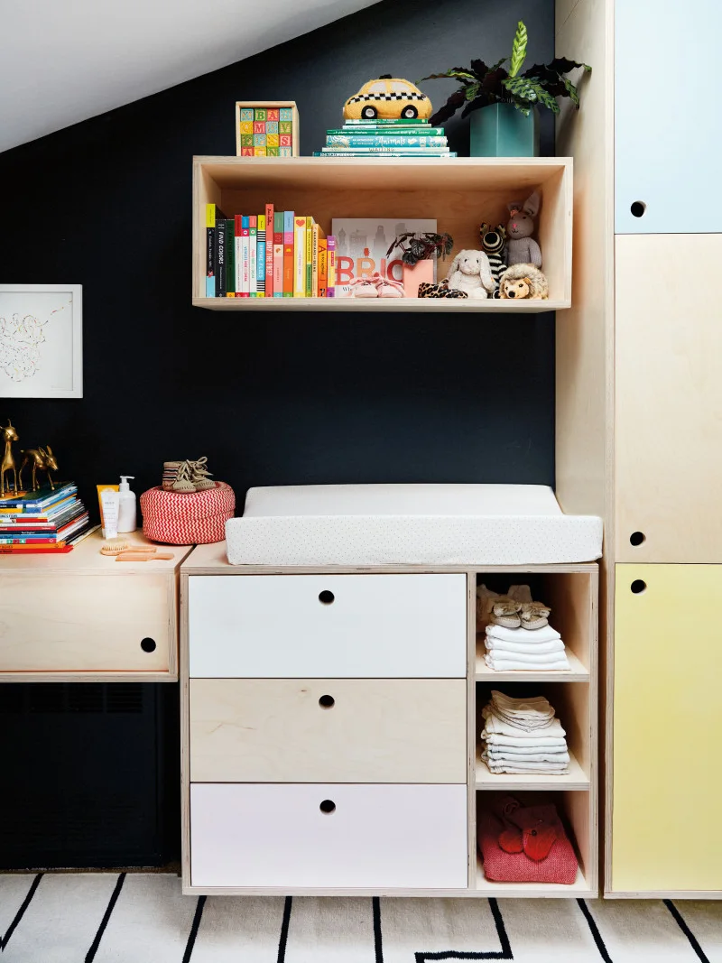A fitted nursery storage wall combines pale colored drawers, open cubbies and a changing pad against a dark wall.