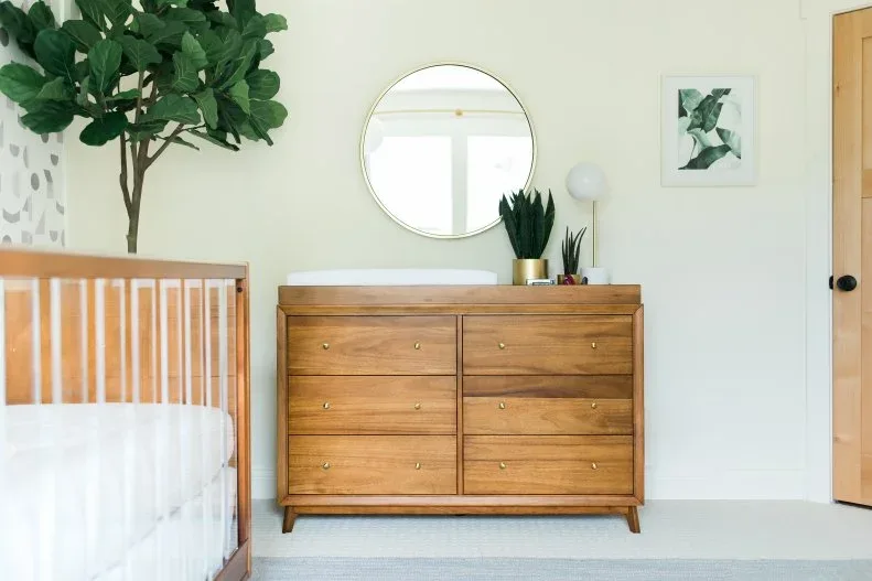 Warm wooden crib and dresser beneath round mirror in pale nursery with broad-leaf plant and smaller plants.
