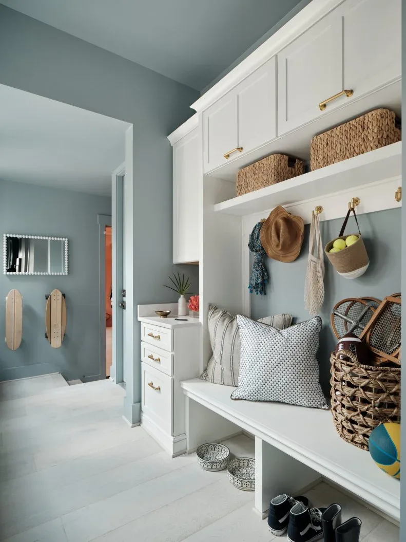 Pale blue and white mudroom with bench, open shoe storage, upper baskets, woven hanging bag and patterned cushions.