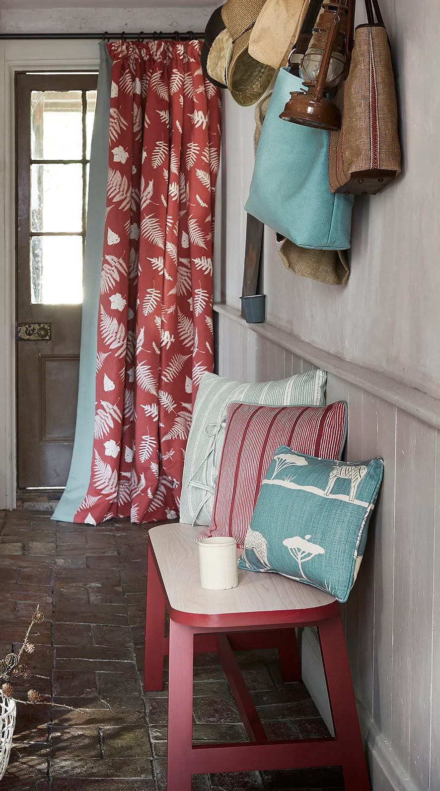 Entrance with a red leaf-patterned door curtain, red bench, blue-green cushions, hanging hats and bags, and pale wall paneling.