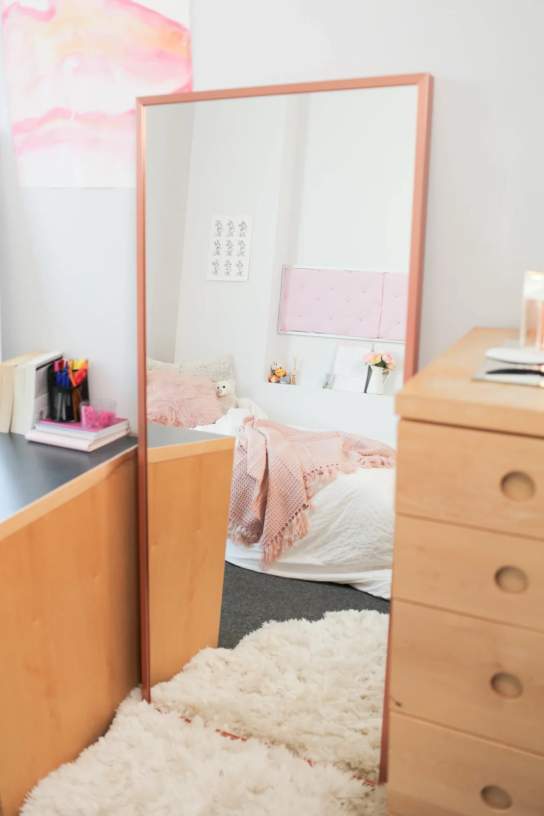 Tall coral-framed mirror between a desk and wood dresser reflecting a pink-and-white student bed