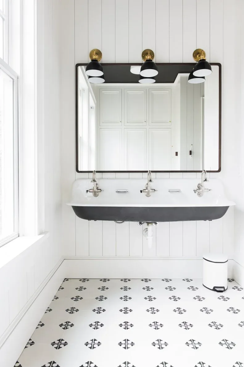 A wall-mounted black-and-white trough basin with two taps sits beneath a broad rectangular mirror and three dark wall lights beside white vertical boards and patterned floor tiles