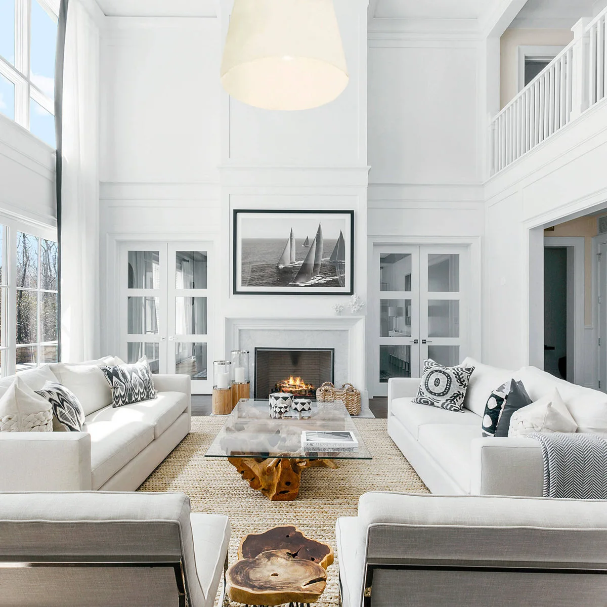 A double-height white living room with pale sofas, a woven rug, wood-based tables and black-and-white cushions.