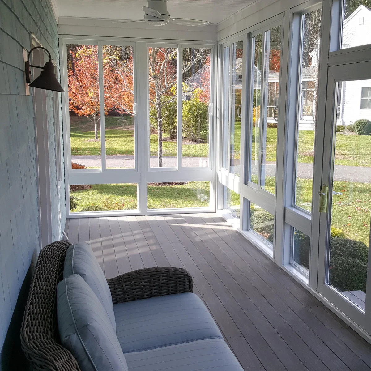 A simple enclosed porch with a wicker chair, gray floorboards, blue-gray siding and tall white-framed opening panels.