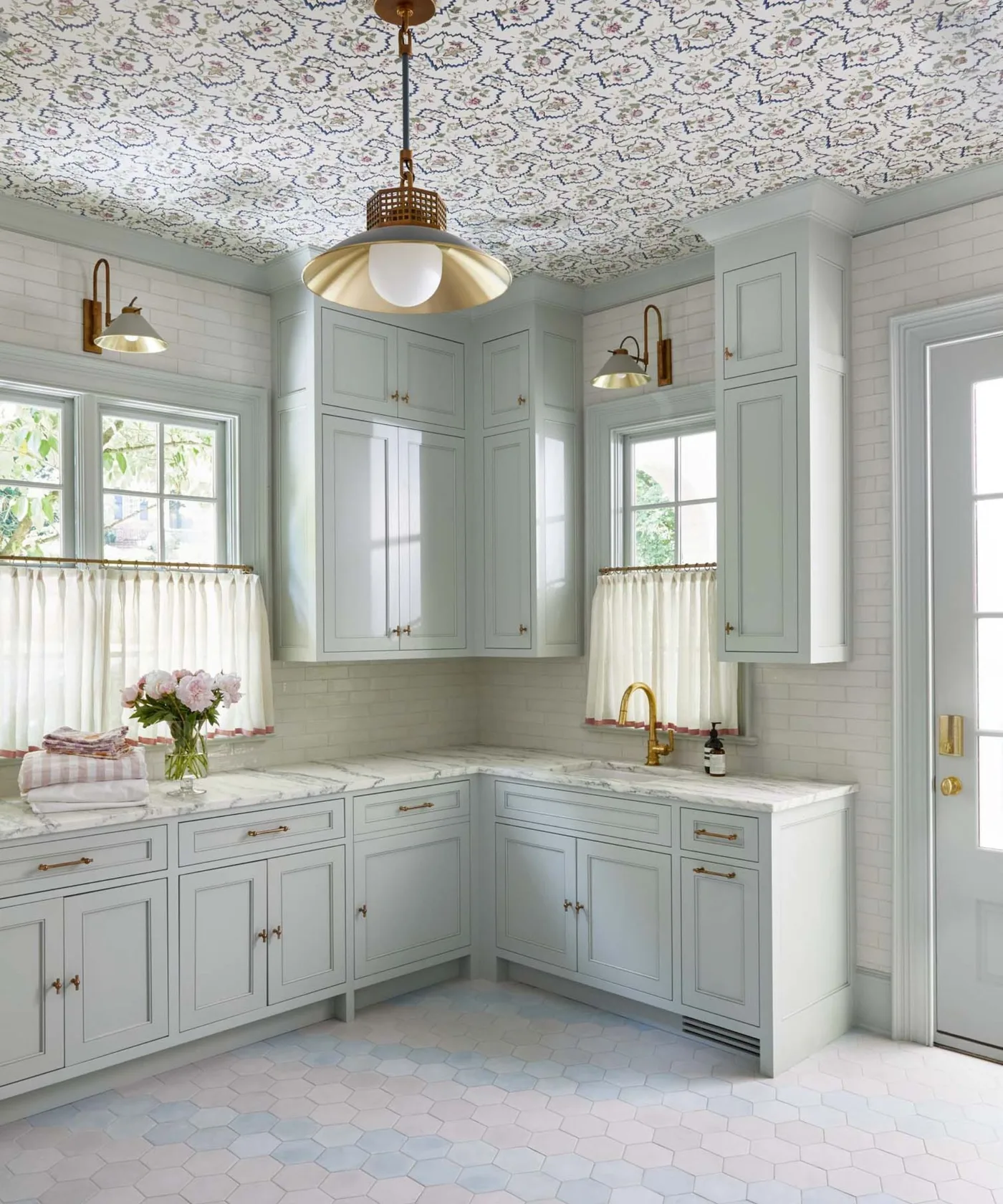 Pale blue utility cupboards with wallpapered ceiling, warm-toned pendant, white tile walls, sink and short window curtains