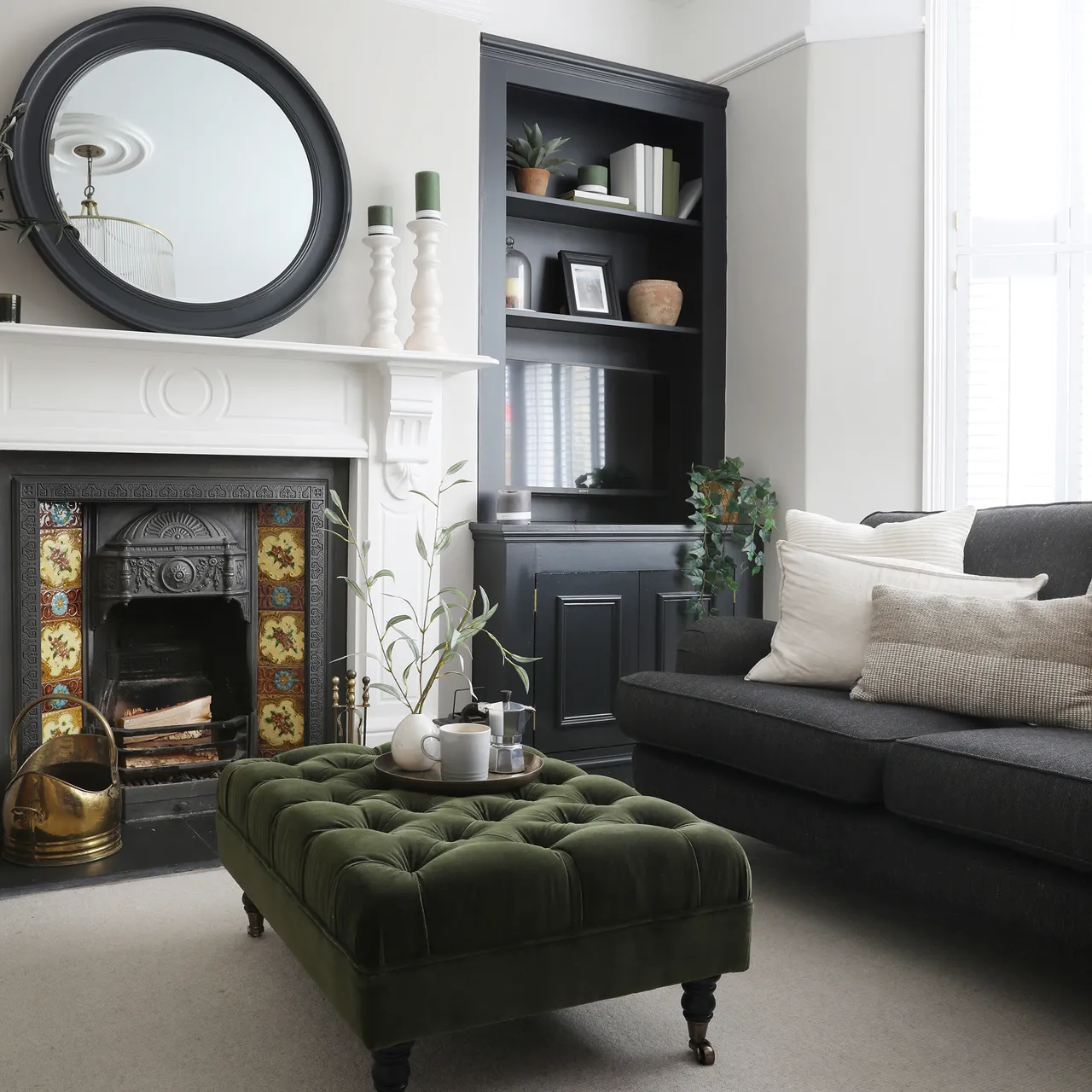 A dark sofa sits beside a white fireplace and dark shelves with a tufted olive-green ottoman