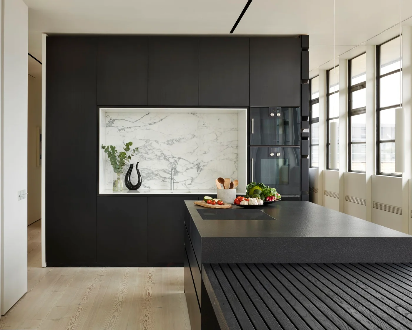 Dark flat kitchen cupboards surround a pale veined sink recess beside tall dark-framed windows and a dark island with a slatted lower section
