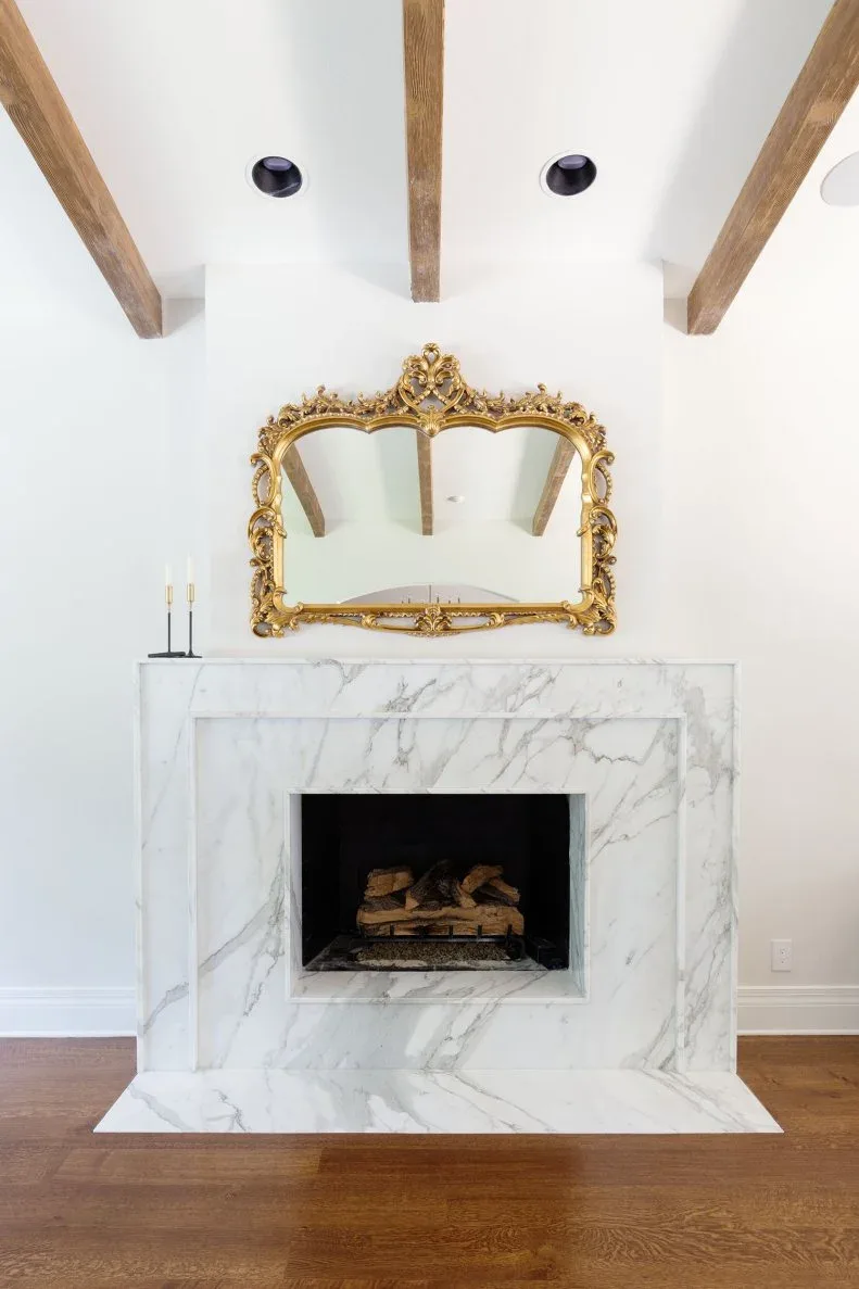 Pale veined fireplace with ornate gold-toned mirror, two slender dark candle holders and wood ceiling beams