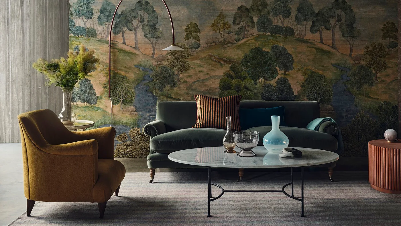A dark green sofa and mustard chair sit before a landscape wallpaper mural with trees and a winding river.