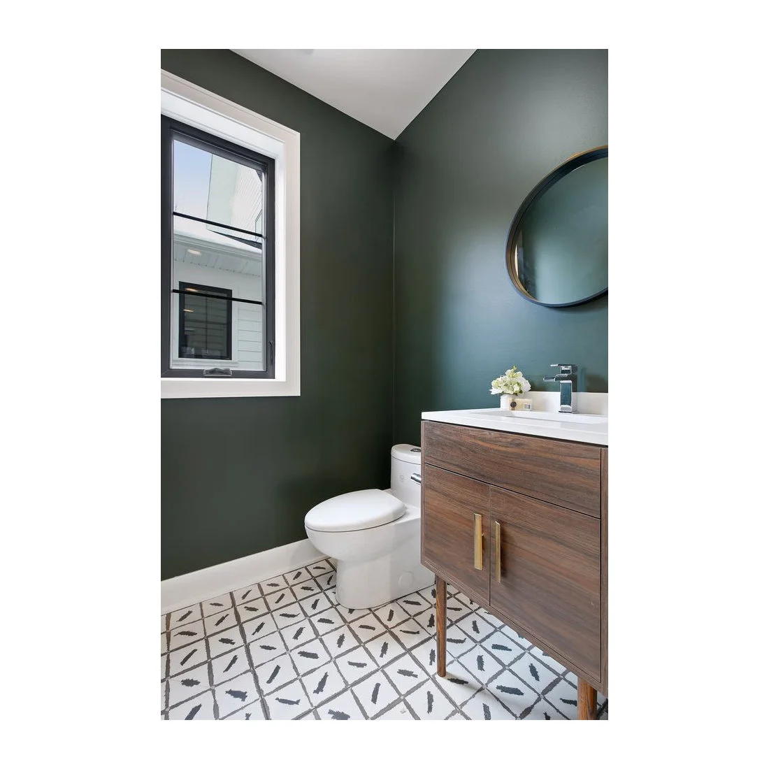 A small powder room has deep green walls, a wood vanity on legs, a round mirror and pale floor tiles with a black graphic line pattern.