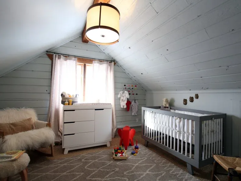 Gray paneled nursery with sloping ceiling, simple crib, pale storage, warm window trim and geometric rug.