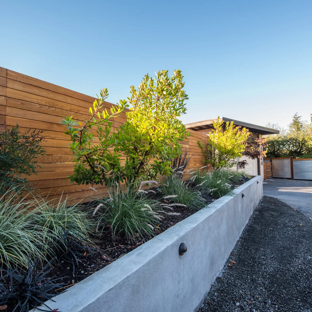 A pale straight retaining wall separates a planted bank and timber fence from a gravel driveway.