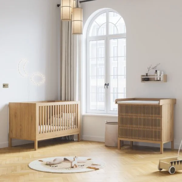 A wooden crib and slatted changing chest stand beside a tall arched window, pale walls and a small patterned round rug