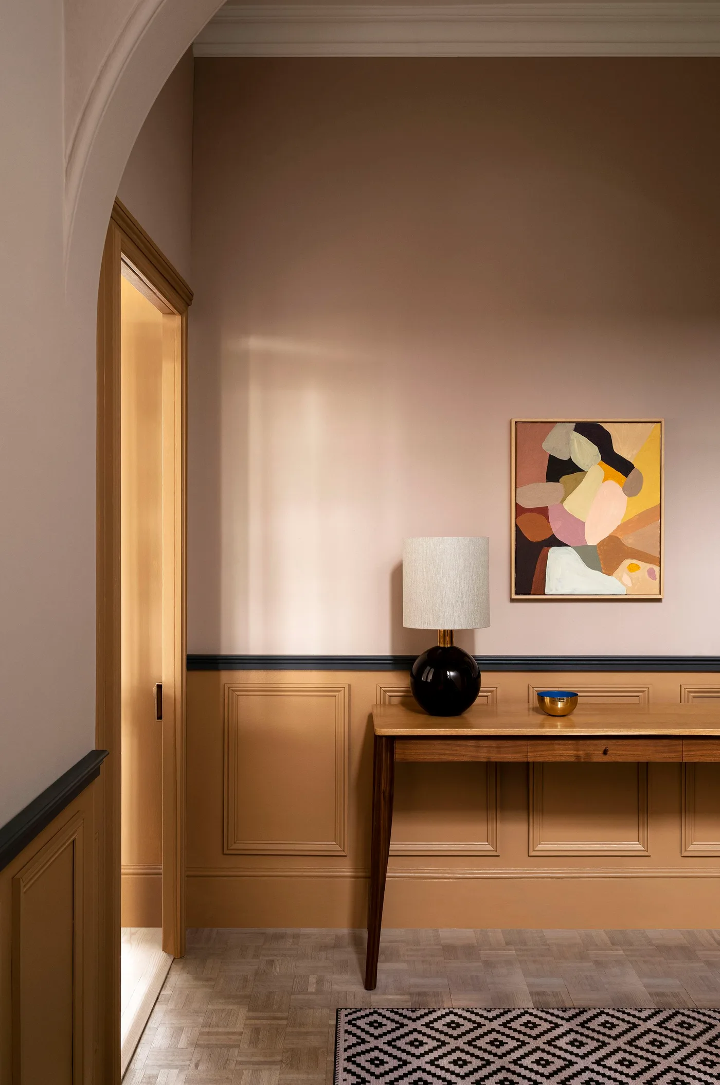 Warm pink hallway with ochre lower paneling, dark trim line, a wooden console and a shaded black lamp.