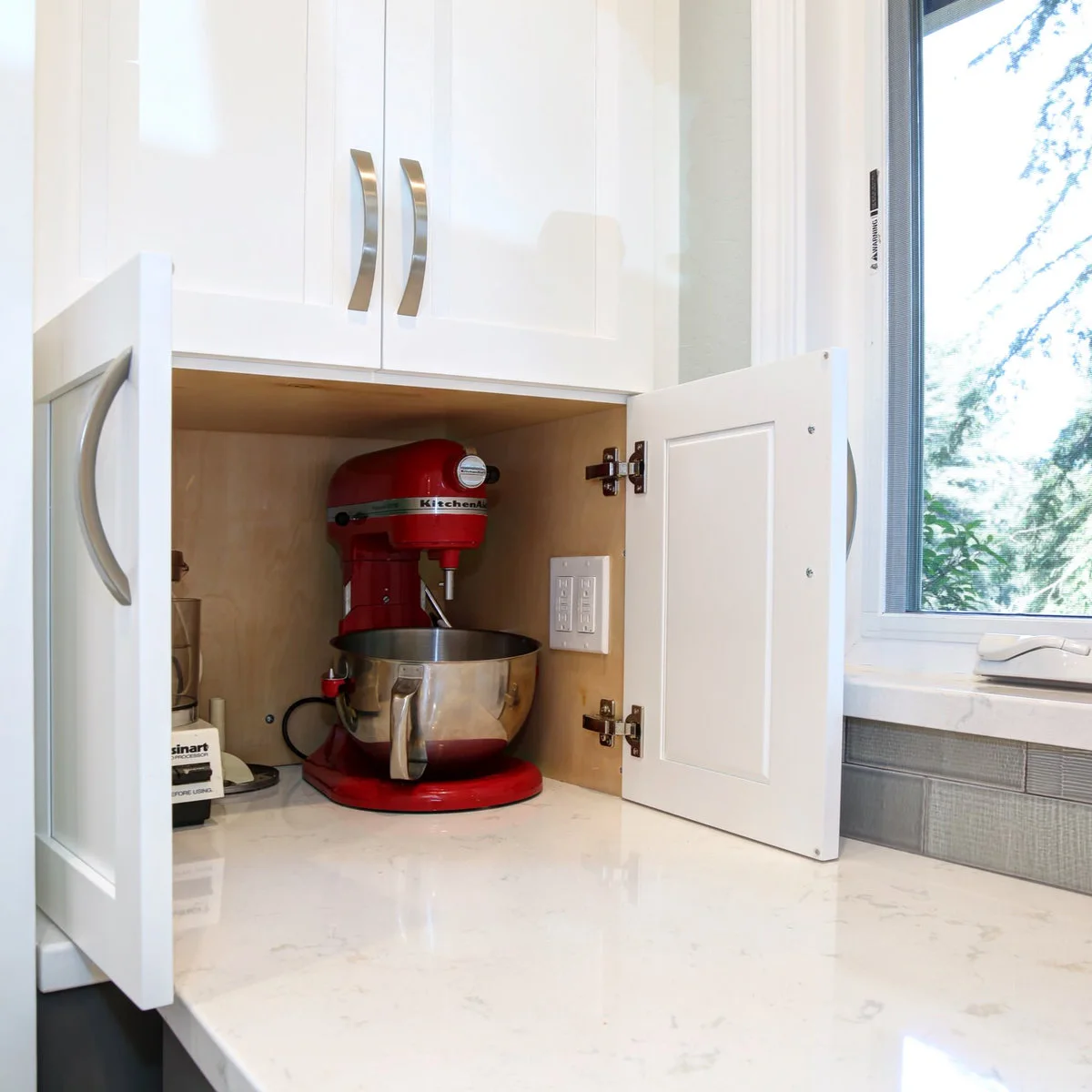 Red stand mixer inside an open white countertop cupboard near a kitchen window
