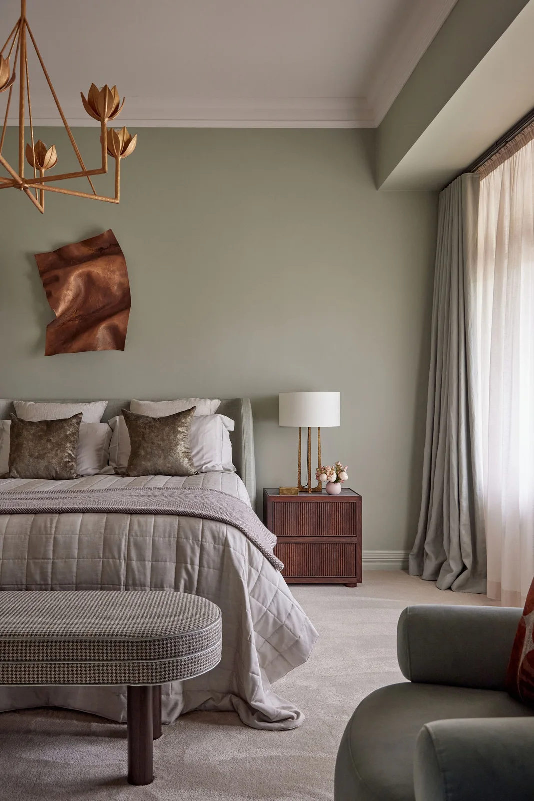 A sage bedroom wall holds a sculptural warm-metal artwork above gray bedding and a dark timber bedside chest.