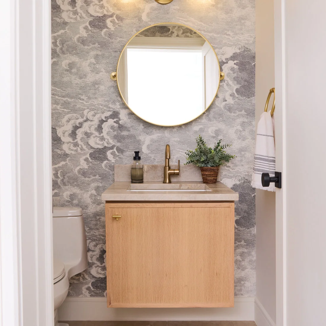 Gray cloud-pattern wallpaper surrounds a small plain wood wall-hung vanity beneath a round warm-metal mirror, beside a white toilet and striped towel.