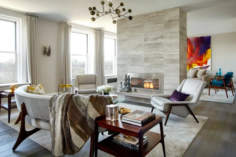 A flat pale fireplace wall with horizontal markings divides a living room with broad windows, light upholstered chairs and a curved sofa