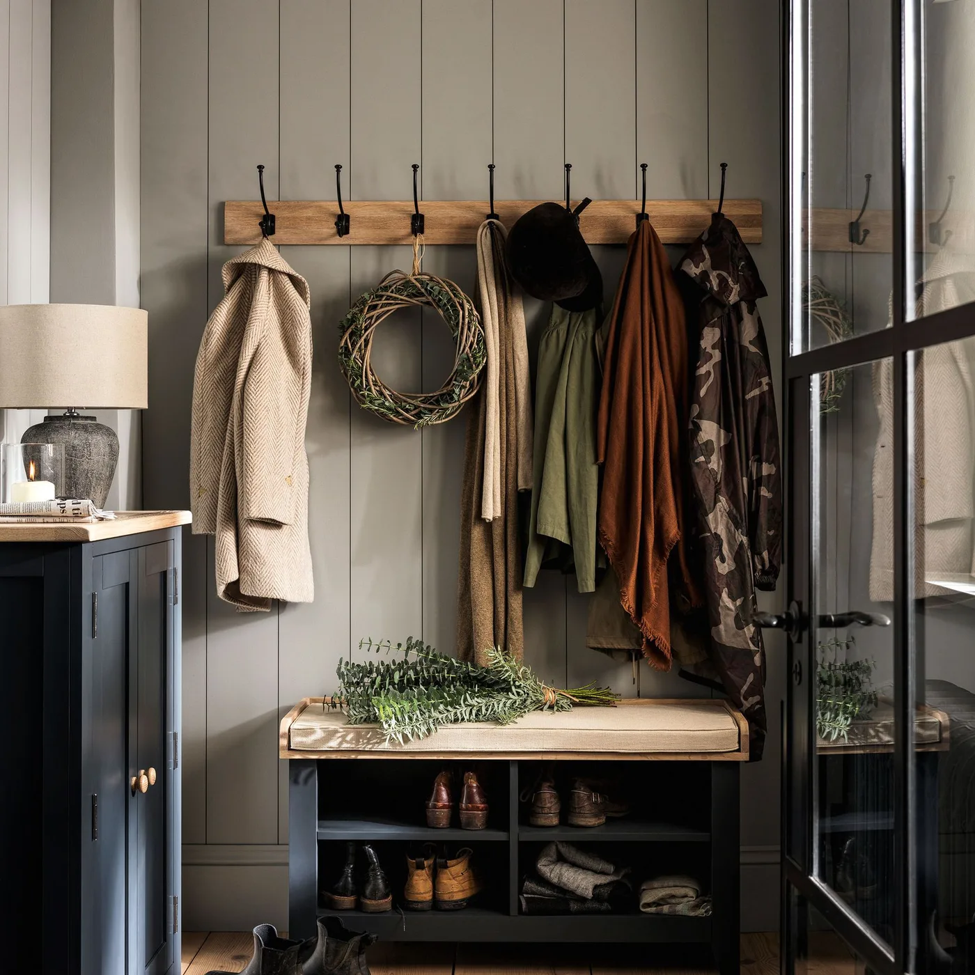 Dark painted entry bench with tan cushion, four open shoe compartments, wood hook rail and pale vertical wall paneling