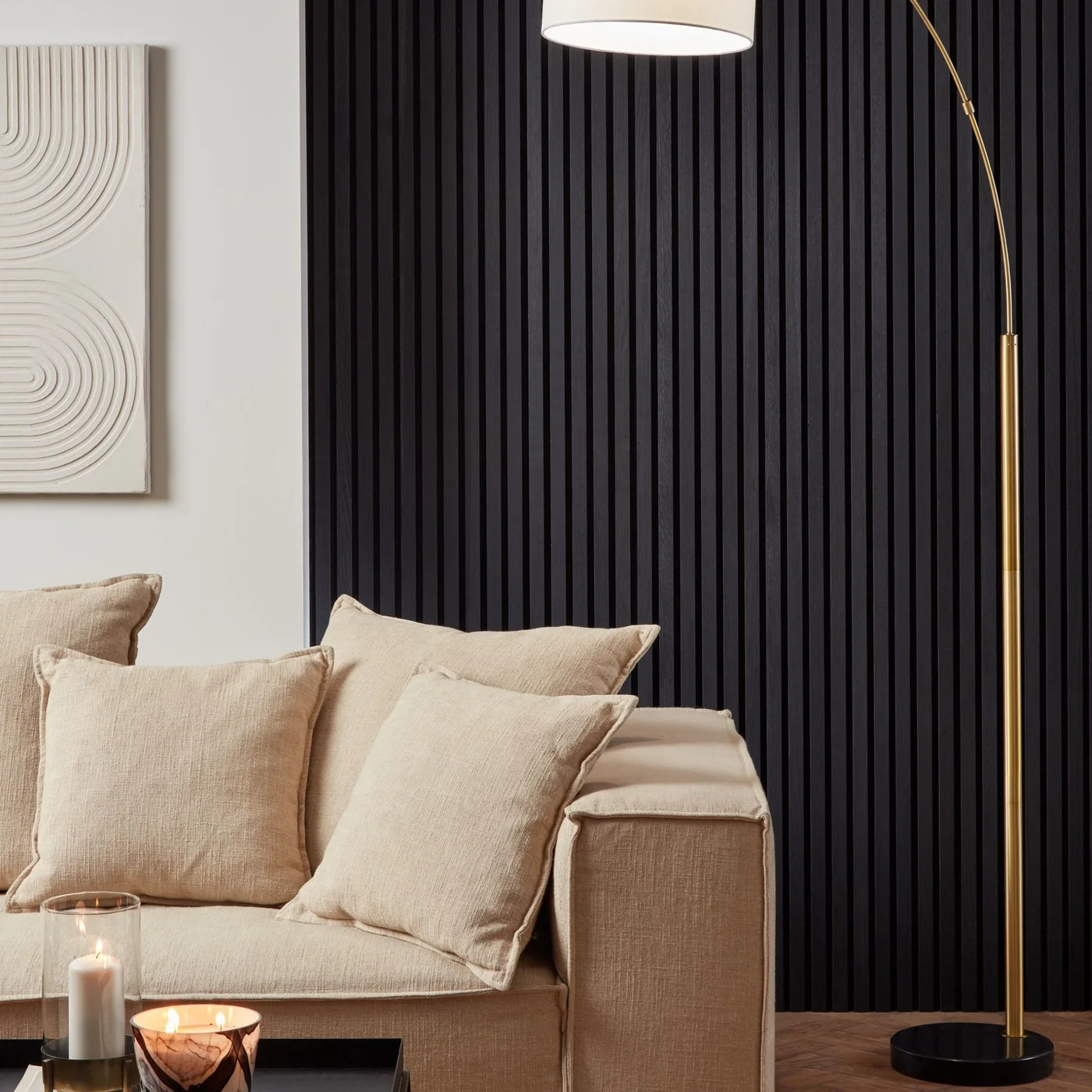 Black vertical wall slats stand behind a beige sofa beside a pale wall and a slim floor lamp