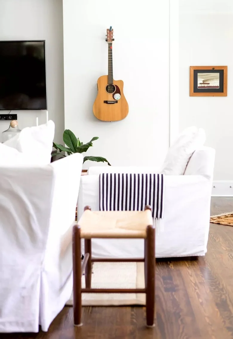 A bright sitting area has white covered seating, a narrow wood stool, striped fabric, a television and an acoustic guitar mounted on the pale wall.