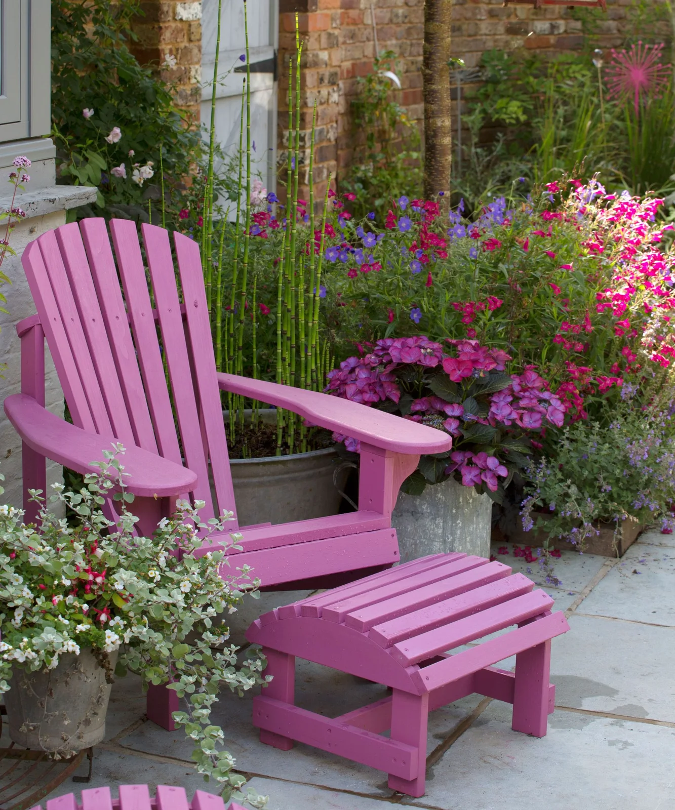 A pink timber lounge chair and footstool sit beside pink and purple container flowers