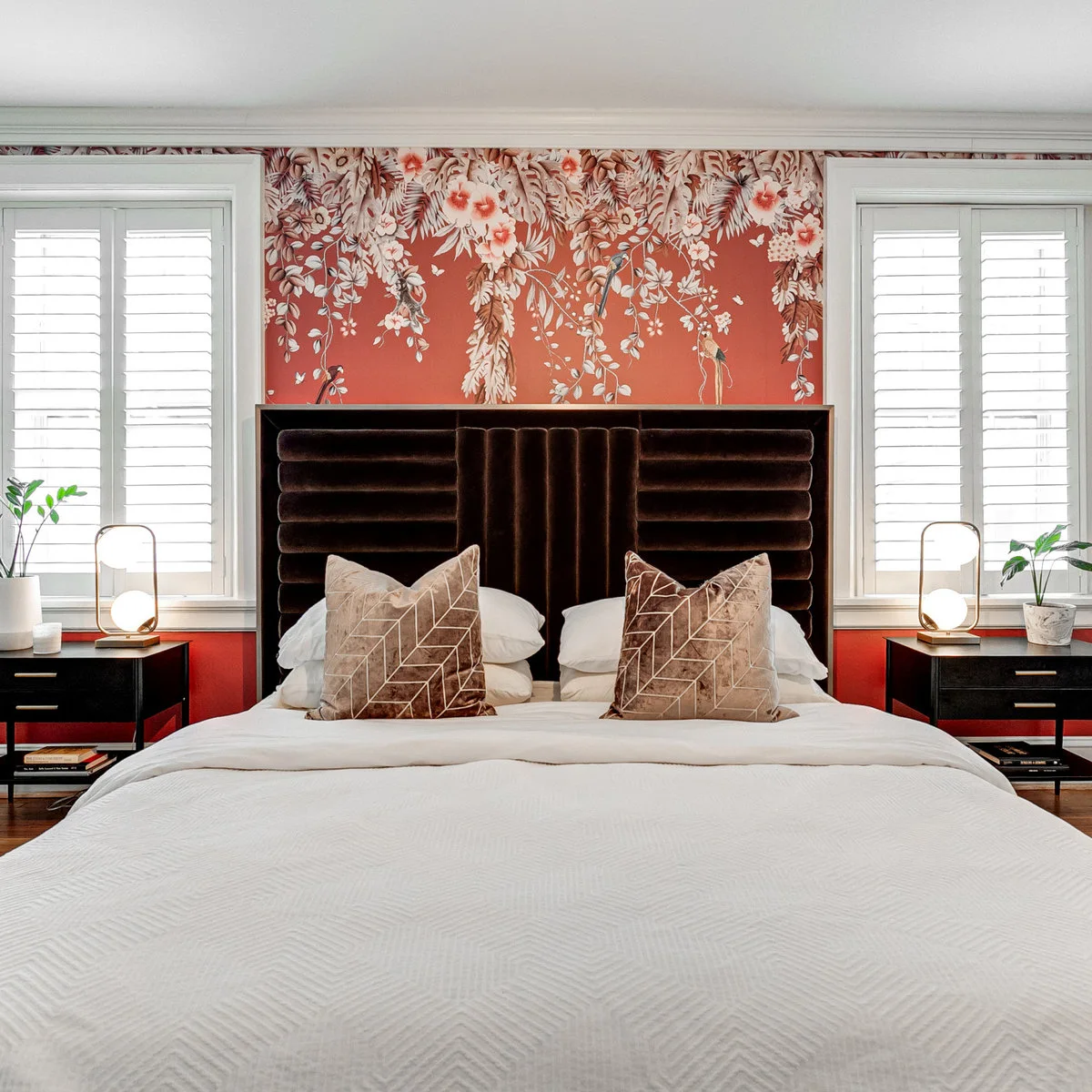 Coral floral wallpaper between white shuttered windows behind a dark channeled headboard with matching bedside tables.