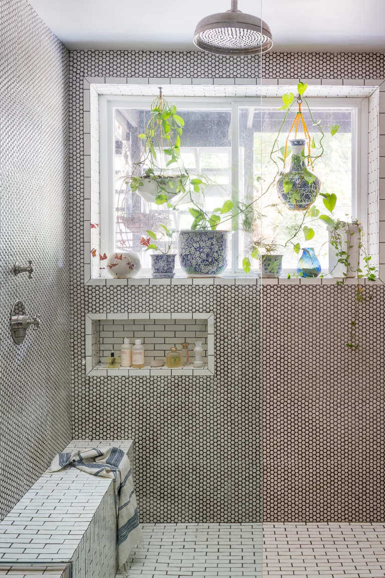 A shower with small black-and-white mosaic walls has a white bench and niche beside a window with plants and hanging pots