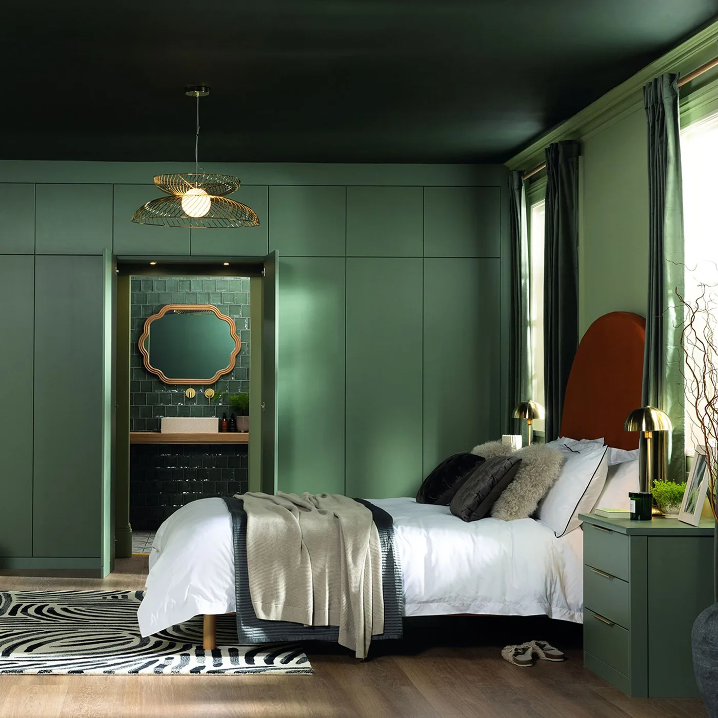 Green built-in cabinetry surrounds a recessed tiled basin area beside a bed with an orange headboard and pale bedding