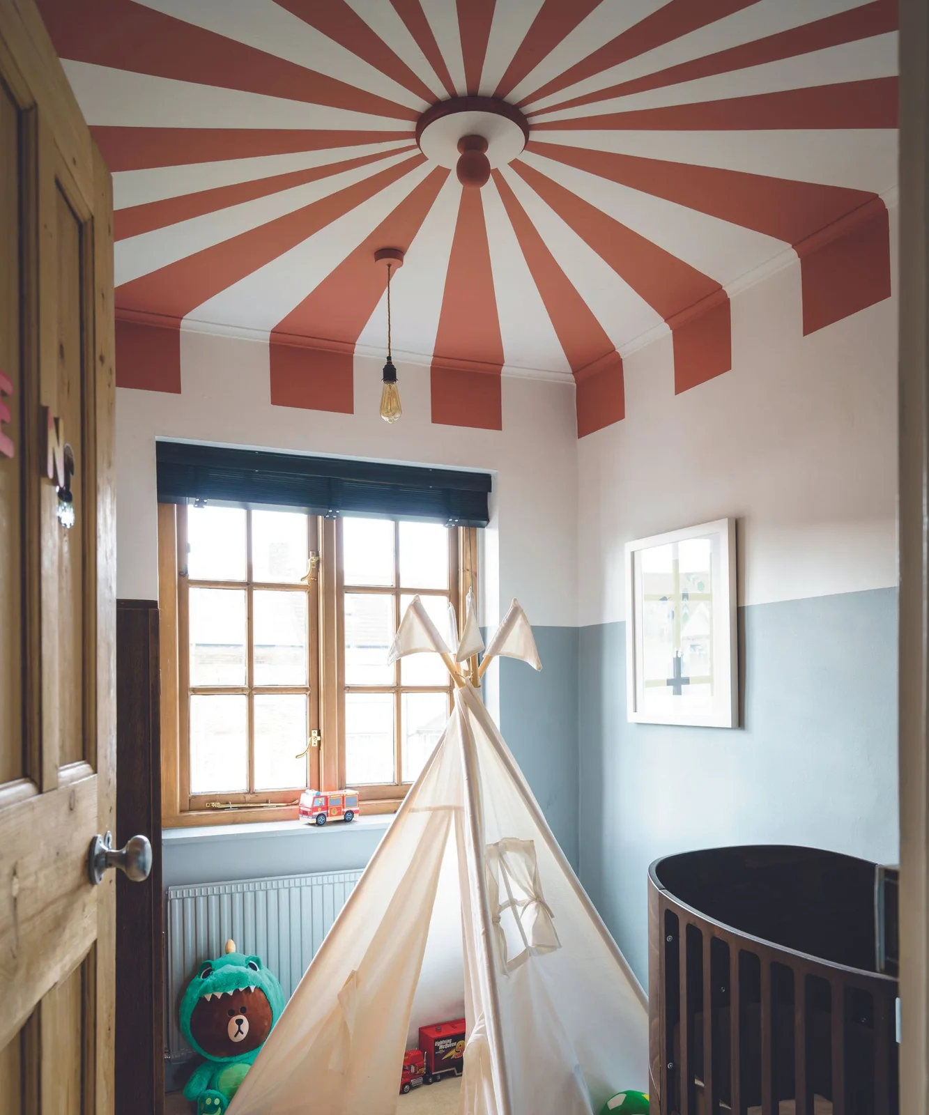 Red-and-white radiating ceiling stripes sit above pale blue lower walls, a wooden window, a play tent and the edge of a dark oval crib