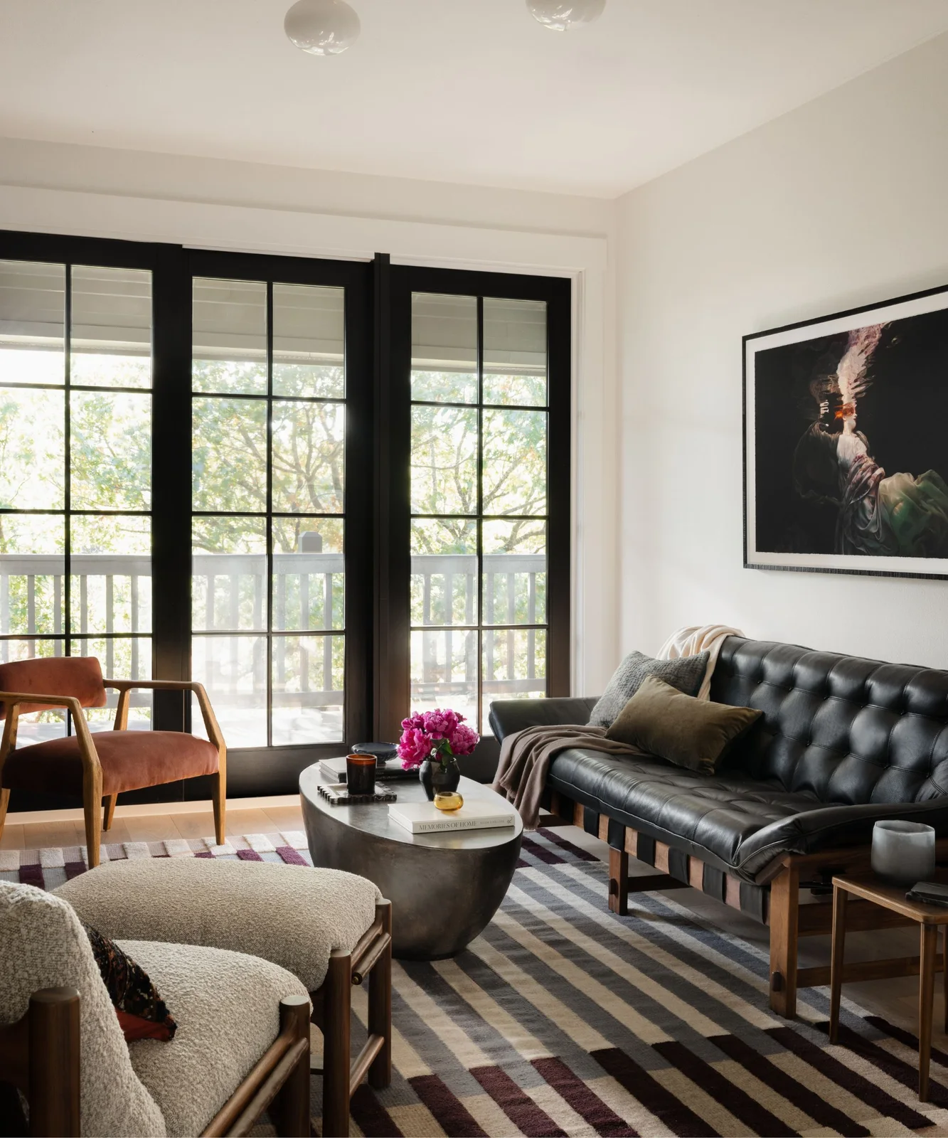 A black sofa with timber legs sits opposite pale timber-framed seating on a striped rug beside tall glazed doors.