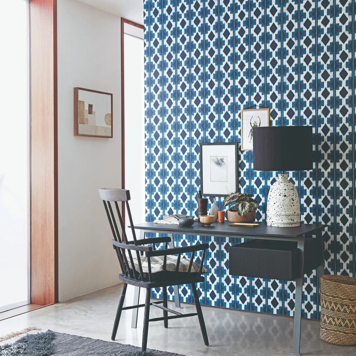 Blue-and-white geometric wallpaper stands behind a dark desk and spindle chair, framed art and a large lamp beside a tall glazed opening.