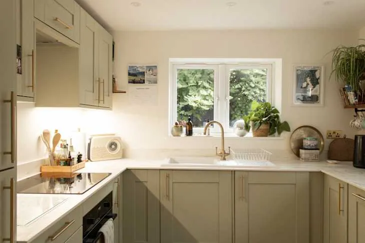 Compact sage-green kitchen with corner worktop, warm timber floor and daylight from a sink window