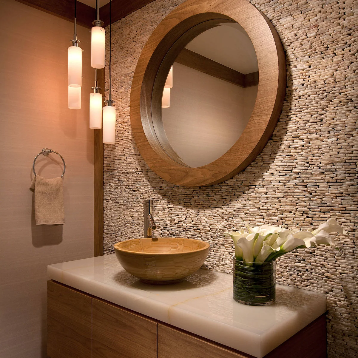 A warm-toned round vessel sink sits below a large round wood-framed mirror against a textured pebble wall.