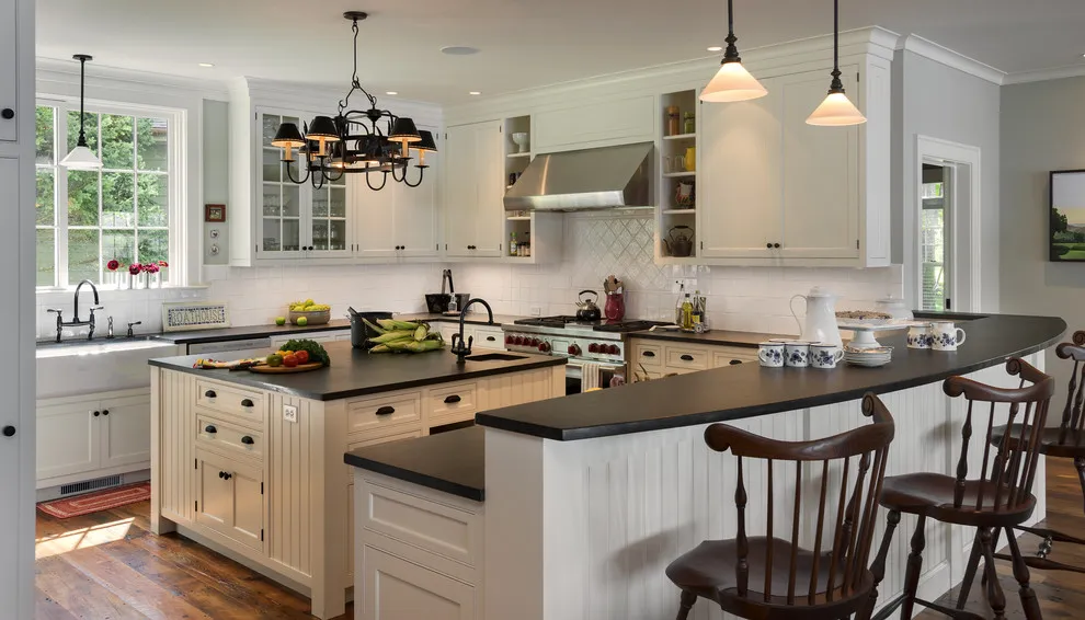 White cabinetry sits beneath dark worktops beside a broad peninsula and timber chairs