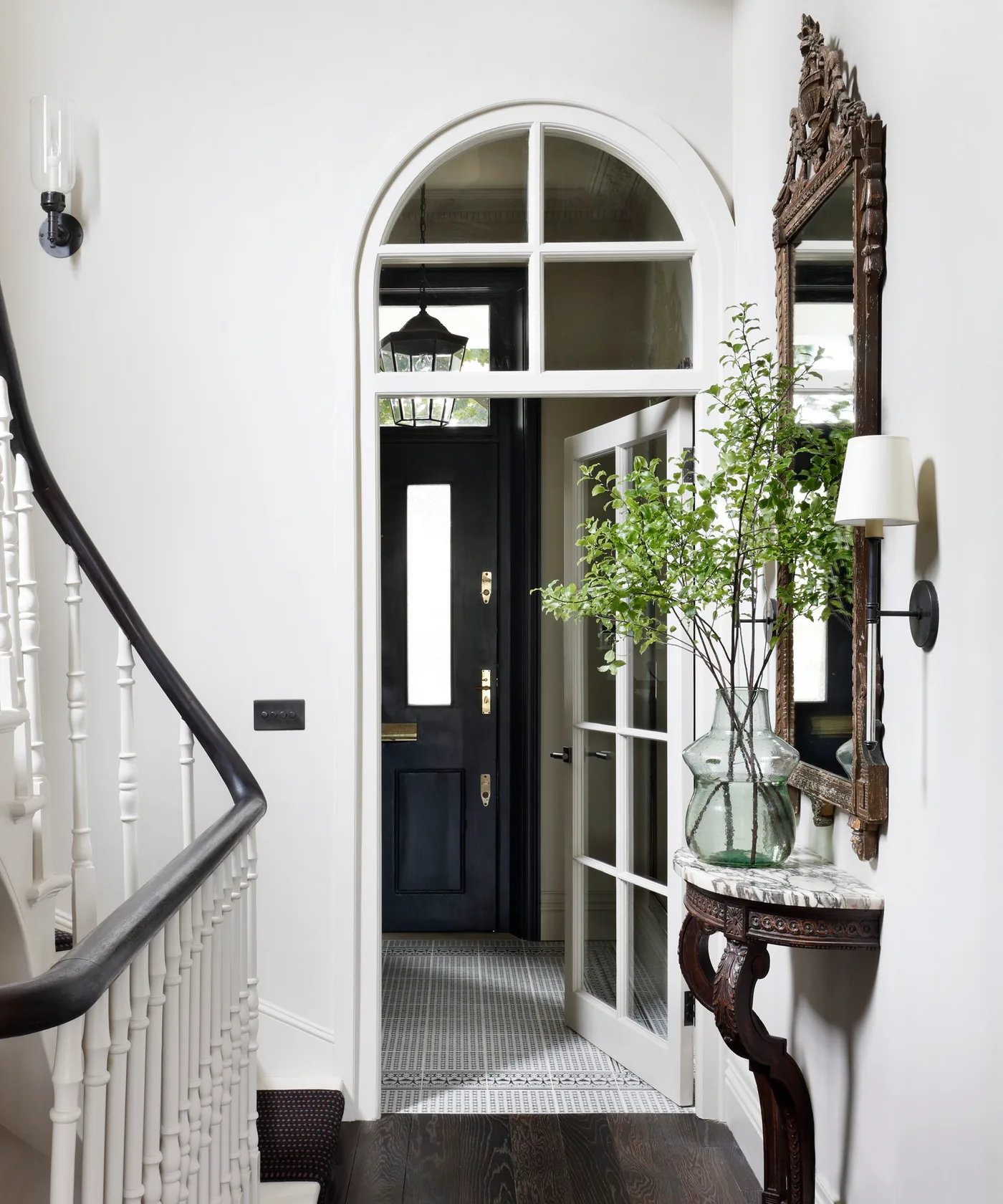 Narrow wall-side console with pale veined top, carved dark supports, tall ornate mirror and leafy branches in a clear vase