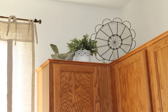 Flower-shaped wire basket, leafy white pot and bird ornament above the corner of warm wood kitchen cabinets.