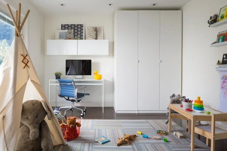 A white room combines tall closed cupboards, a computer desk with a blue chair, a small wood play table, a fabric tent and a striped floor rug.